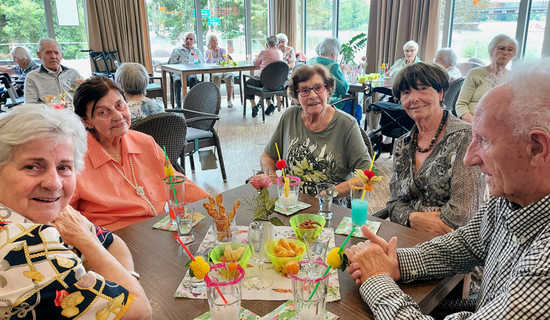 Bewohner*innen und Mieter*innen aus dem Service-Wohnen der ProCurand Seniorenresidenz Havelpalais sitzen in der festlich mit bunten Girlanden geschmückten Cafeteria im Kreis, stoßen fröhlich mit ihren Cocktails an und genießen den gemütlichen Nachmittag.