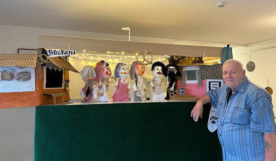 Ein Bewohner des ProCurand Pflegestifts Pfarrer Lukas in Cham steht an einer Werkbank, auf der mehrere selbst gebastelte Handpuppen arrangiert sind.