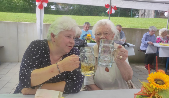 Zwei Seniorinnen stoßen im Garten des ProCurand Seniorenzentrums Am Herzogschloss mit Biergläsern an und genießen den lauen Sommerabend.