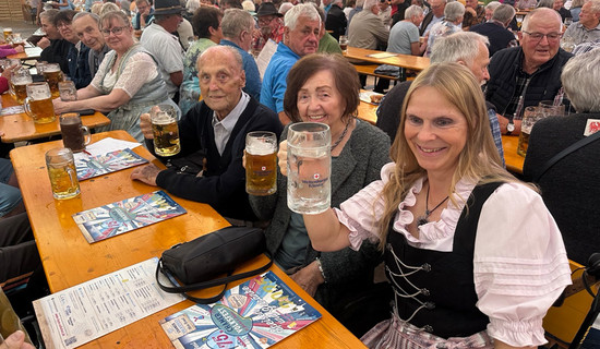 Die Bewohner*innen und eine Mitarbeiterin des ProCurand Pflegestifts Pfarrer Lukas sitzen am Tisch zusammen, lachen und stoßen gemeinsam mit Bierkrügen auf einen fröhlichen Nachmittag beim Chamer Volksfest an.