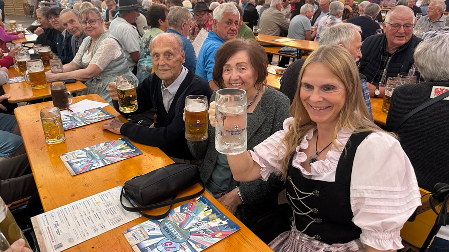 Die Bewohner*innen und eine Mitarbeiterin des ProCurand Pflegestifts Pfarrer Lukas sitzen am Tisch zusammen, lachen und stoßen gemeinsam mit Bierkrügen auf einen fröhlichen Nachmittag beim Chamer Volksfest an.