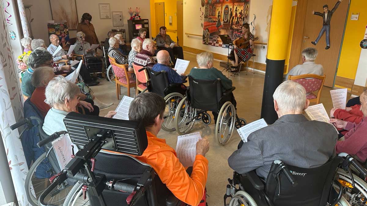 Bewohner*innen sitzen gemeinsam im Raum, verfolgen aufmerksam das Geschehen und nehmen aktiv am musikalischen Programm teil.