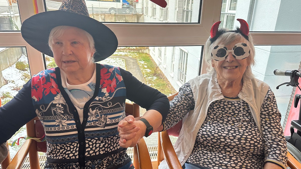 Zwei Bewohnerinnen sitzen nebeneinander, lachen und halten sich in bunten Kost&uuml;men fr&ouml;hlich an den H&auml;nden.